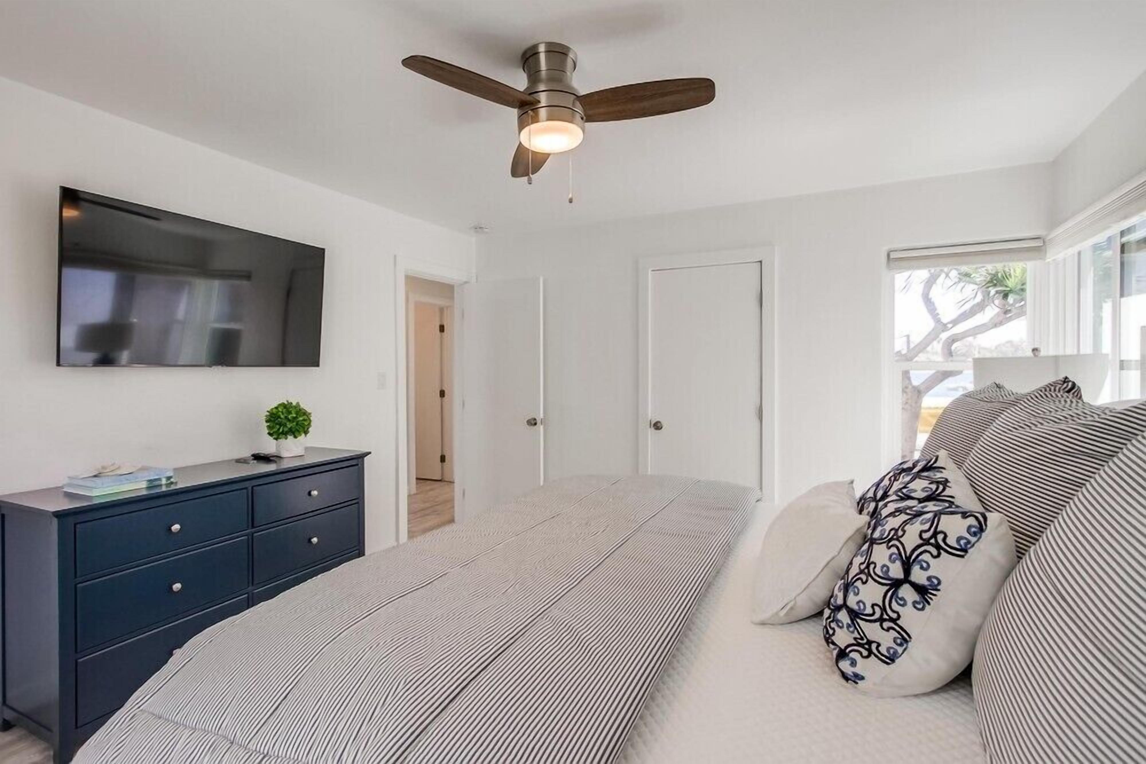 Un dormitorio con cama, tocador, ventilador de techo, televisor y ventana.
