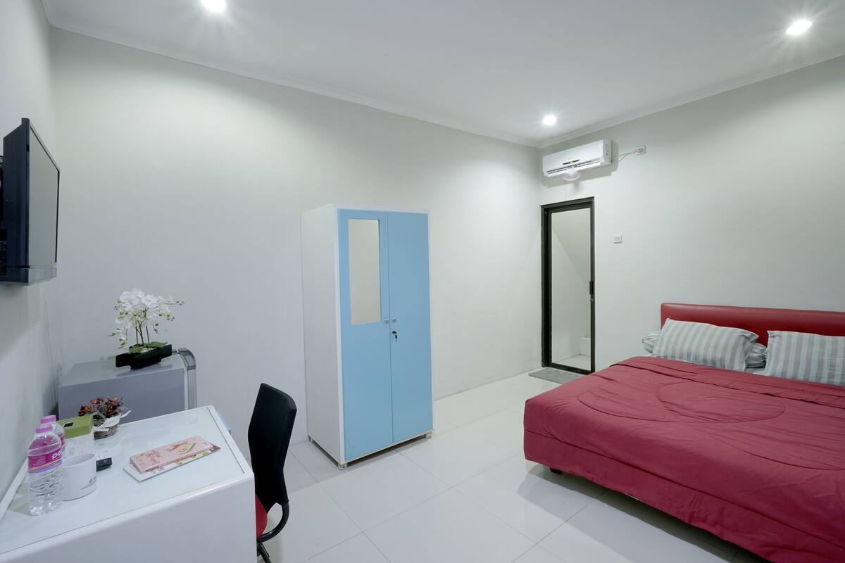 Quarto com cama vermelha, guarda-roupa azul, escrivaninha com cadeira e uma pequena mesa com planta e garrafa de água.