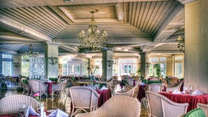 Une salle à manger spacieuse, dotée de chaises et de tables en osier, de grandes fenêtres et d’un plafond haut orné d’un lustre.