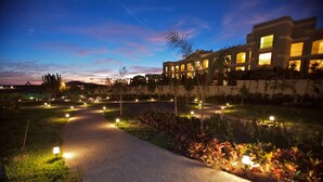 Un chemin bien éclairé mène à un bâtiment doté de nombreuses fenêtres et à un jardin aménagé, agrémenté de petites lumières.