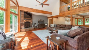 A spacious living room with a fireplace, wooden beams, a balcony, and a large window. - Lake Days & Game Nights w/ Hot Tub (Oakland)