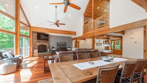 A spacious living room with a wooden dining table, comfortable seating, a fireplace, and a balcony with a wooden railing. - Lake Days & Game Nights w/ Hot Tub (Oakland)