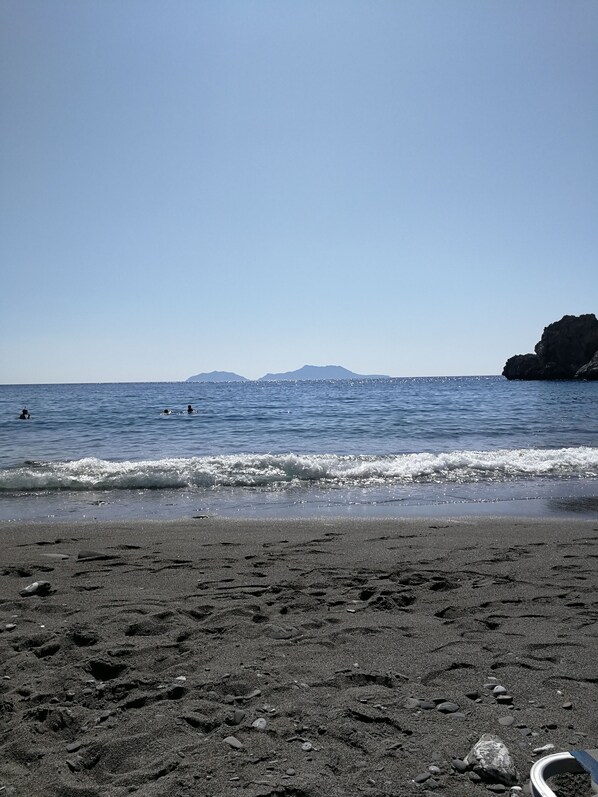A beach with dark sand, waves, and distant mountains.