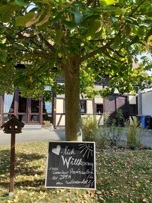 Un cartello di benvenuto sotto un albero, un mangiatoia per uccelli e un edificio con ampie finestre sullo sfondo.