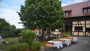 Un espace de restauration en plein air, avec des tables et des bancs, entouré de verdure et bordé d’un bâtiment dont le toit est recouvert de tuiles.