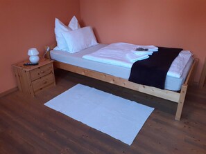 Una cama individual con estructura de madera, ropa de cama blanca y un plaid negro. Hay una mesita de noche de madera con una lámpara y un control remoto, y una alfombra blanca en el suelo.