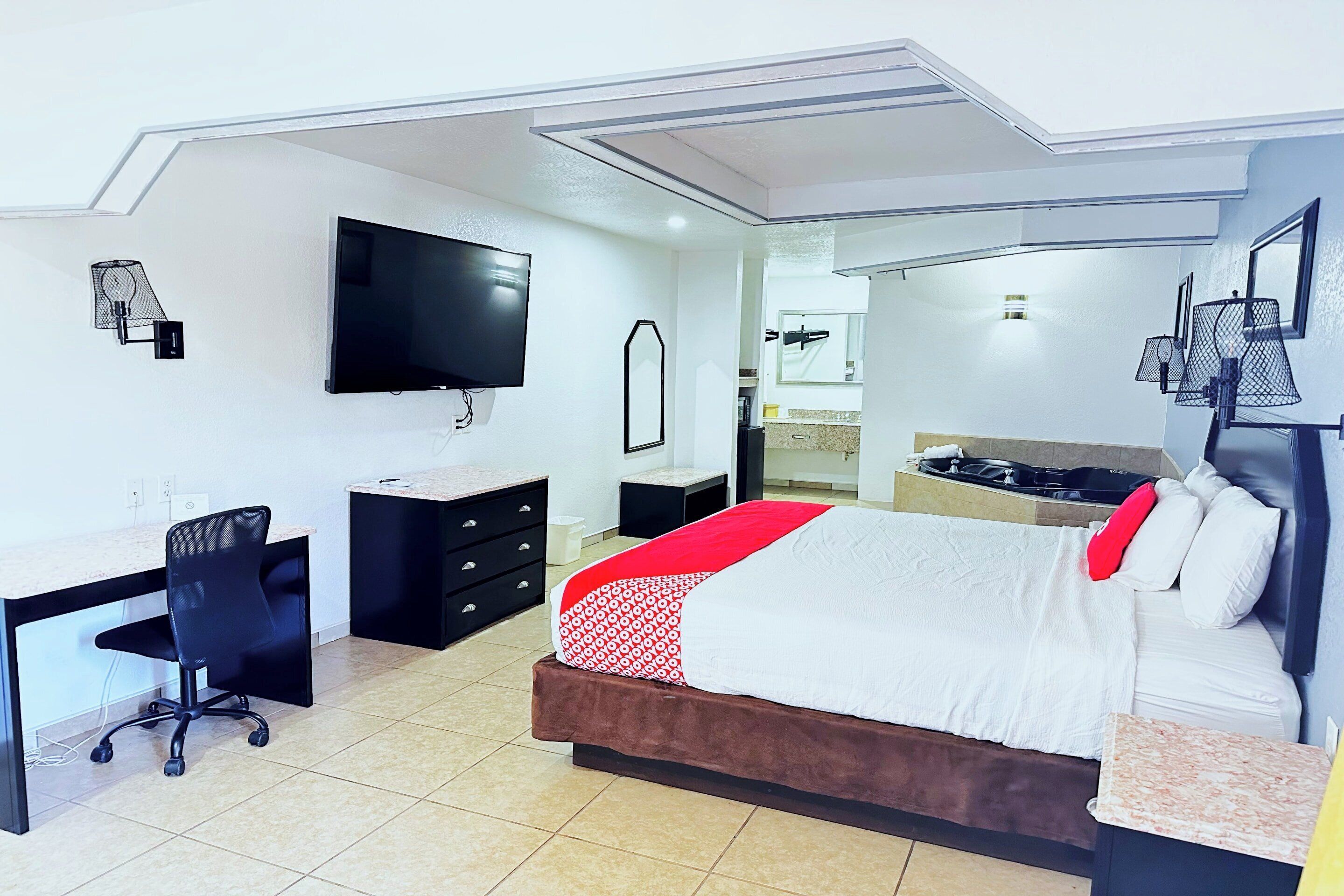 A modern hotel room with a bed, desk, chair, TV, and a bathroom visible through a glass partition.