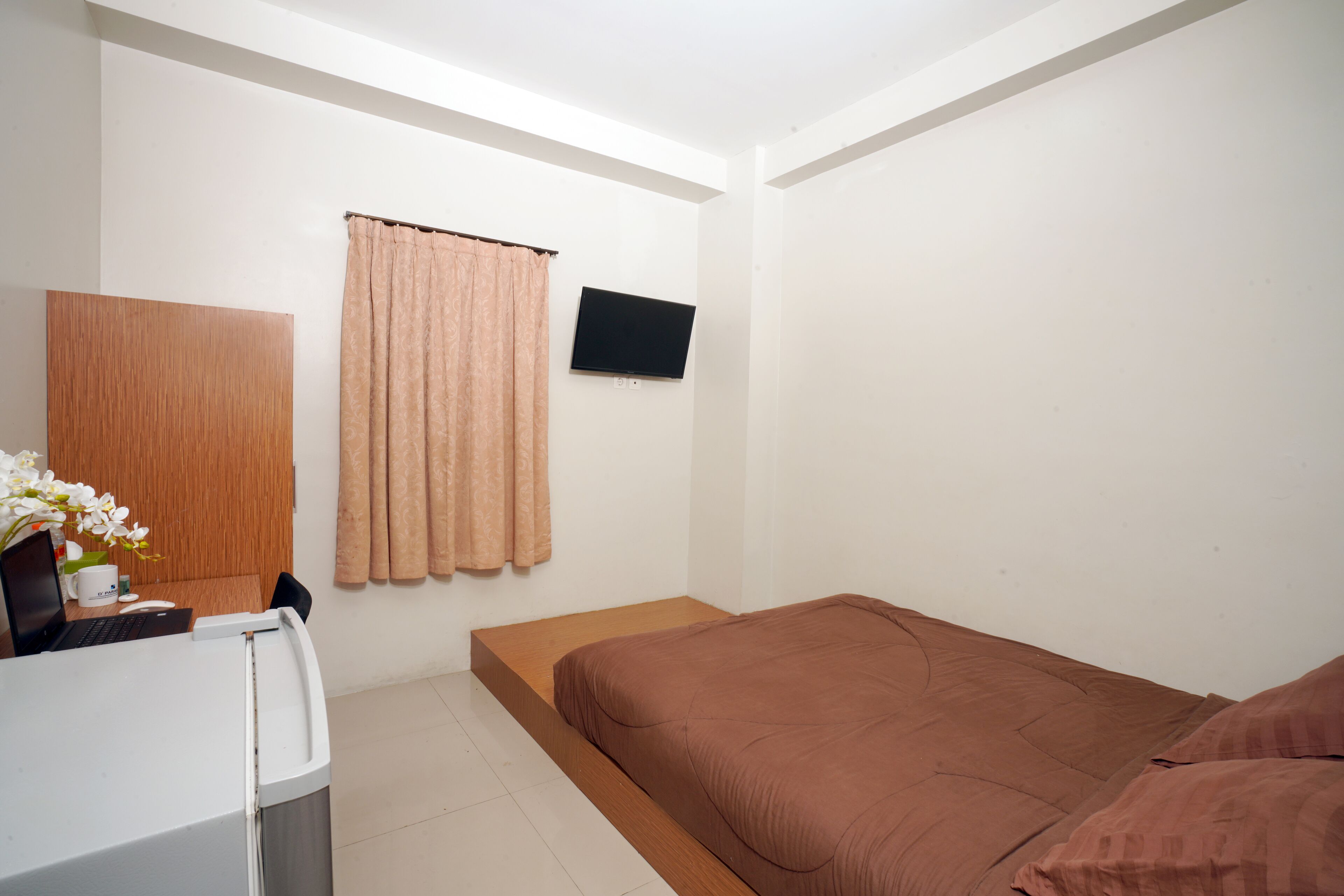 A bedroom with a bed, a desk with a laptop, a television mounted on the wall, and a curtain.