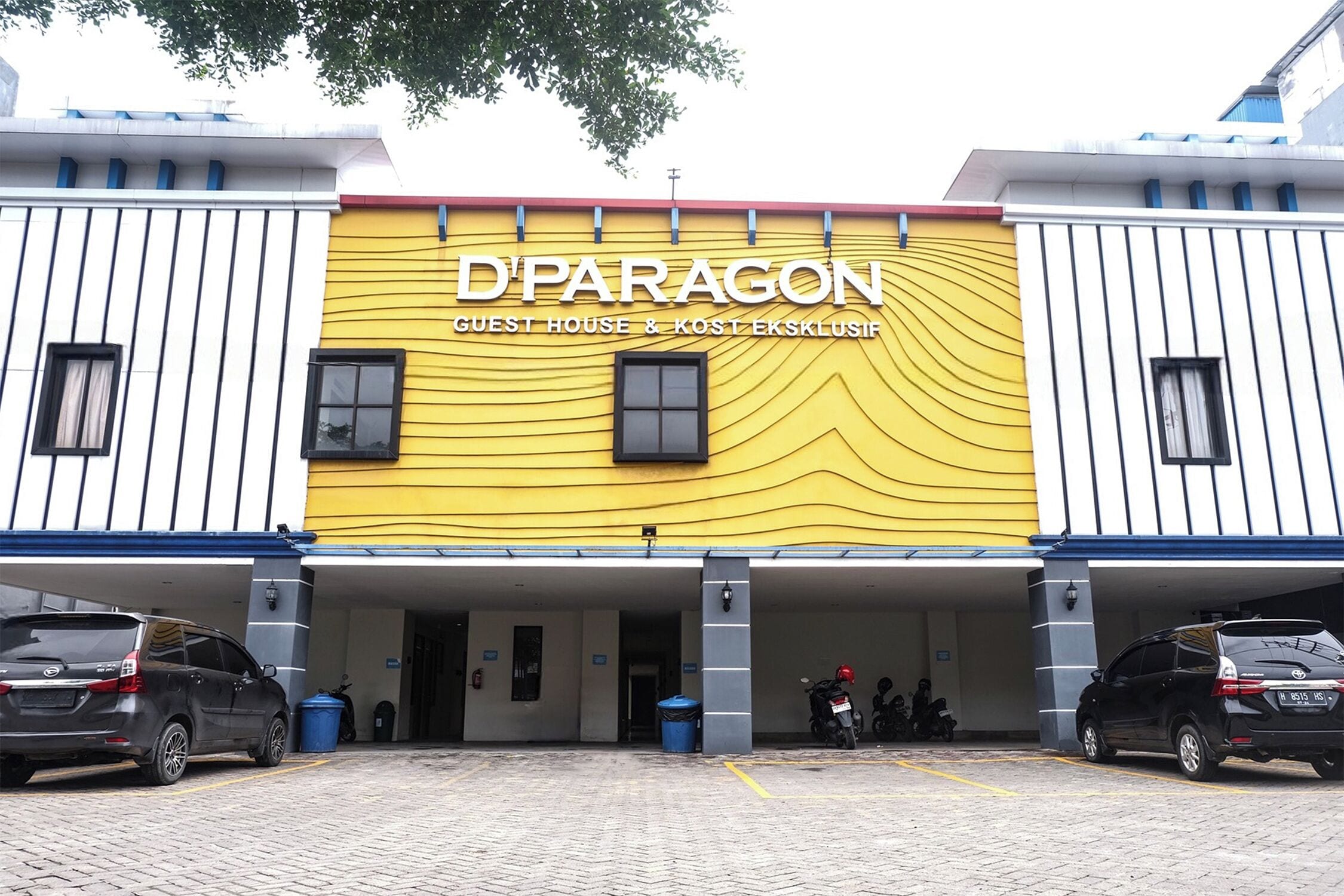Een gebouw met een gele gevel en de tekst 'D'PARAGON GUEST HOUSE & KOST EKSKLUSIF' duidelijk zichtbaar.