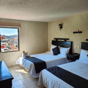 Habitación de hotel con dos camas, una ventana con vista a la ciudad, una mesita de noche y una lámpara de pared.