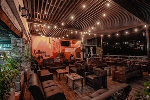 Un espacio al aire libre con mobiliario de madera y luces colgantes, que presenta un mural y una pantalla de televisión.