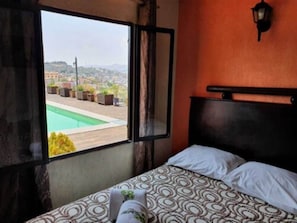 Quarto com cama, janela com vista para uma piscina e luminária na parede.