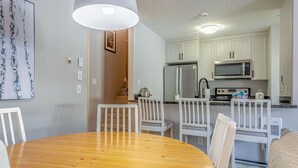 A compact kitchen with white cabinets, a microwave, and a refrigerator. There is a dining area with a wooden table and white chairs. - Crystal Forest #29 by Bear Country (Sun Peaks)