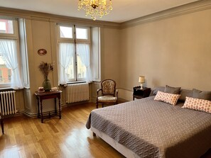 A bedroom with a large bed, a chair, a table with a lamp, and a window with curtains. - Magnifique Appartement Style Maison de Famille (Mouthe)