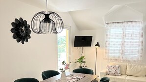 Un salon moderne avec une table à manger en bois, des chaises vertes, un luminaire suspendu noir, une décoration murale noire, un téléviseur à écran plat, une lampe sur pied, un canapé avec des coussins à motifs floraux et une fenêtre avec des rideaux à motifs floraux.