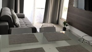 Un salon moderne avec une table à manger en verre, un canapé gris et de grandes fenêtres avec des rideaux.