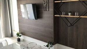 Un salon moderne avec un téléviseur à écran plat fixé sur un mur recouvert de panneaux en bois foncé, une table basse en verre avec un pied en métal et une étagère au design géométrique agrémentée d’objets décoratifs.