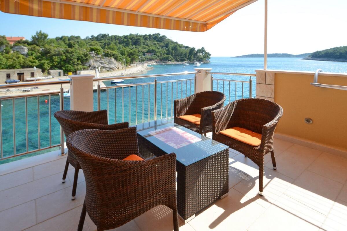 A balcony with wicker furniture, a glass table, and a view of the sea and greenery.