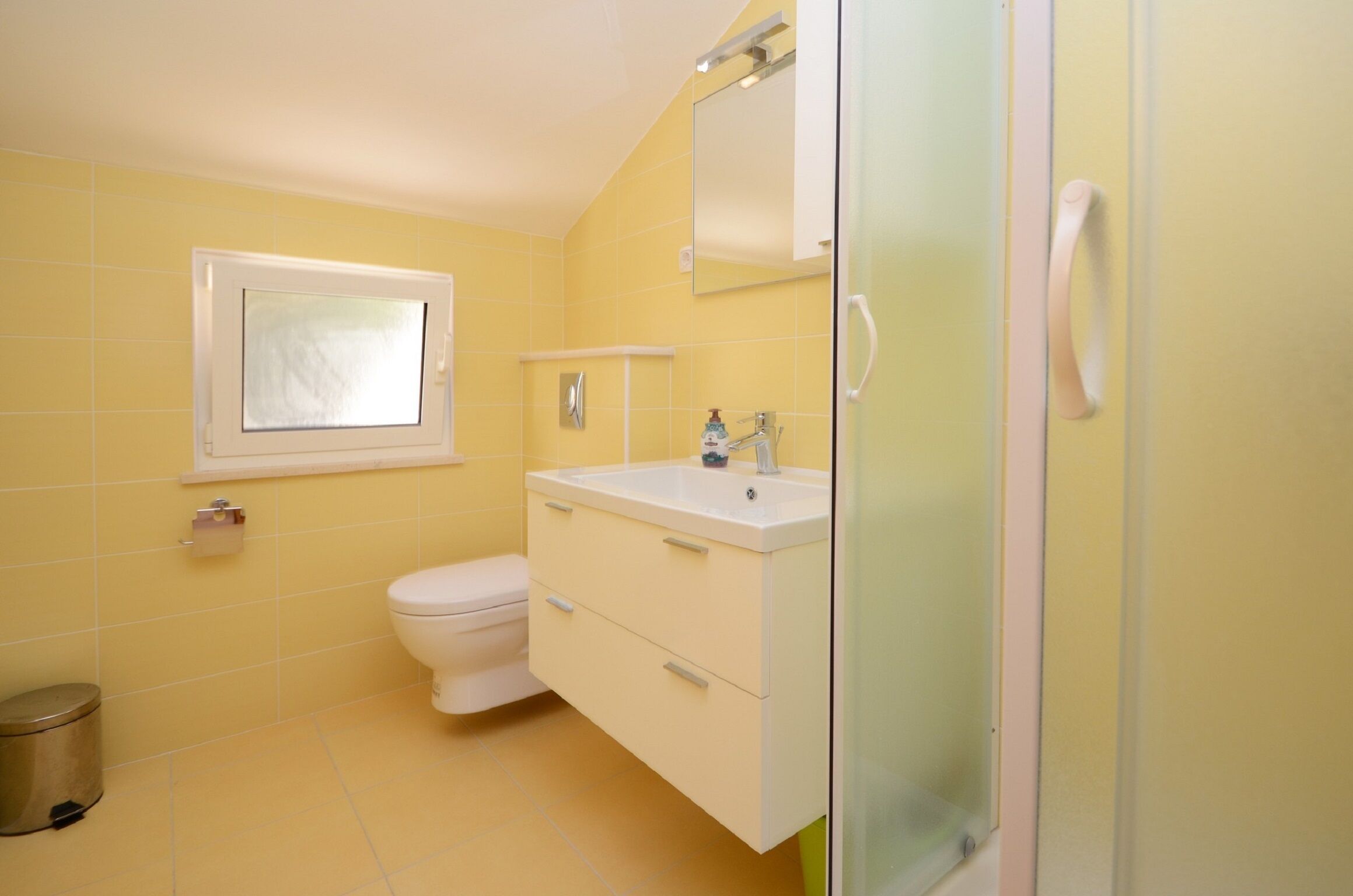 Un baño con paredes amarillas, un inodoro blanco, un lavabo con espejo y una zona de ducha.