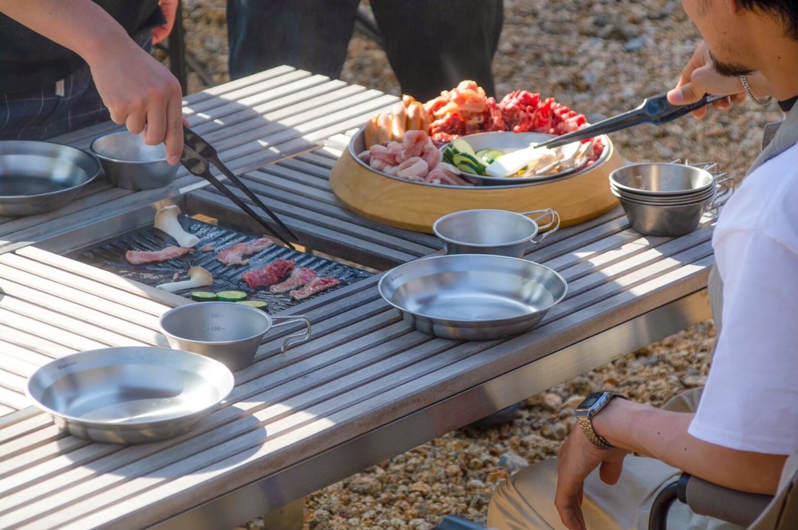 Ein Grill mit verschiedenen Fleischsorten und Gemüsesorten, umgeben von Metallschüsseln und Besteck.