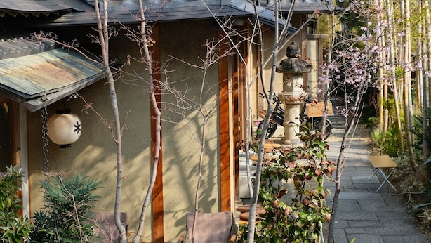 Un bâtiment traditionnel japonais, doté d’un toit recouvert de tuiles, d’une lanterne en pierre et d’un petit jardin agrémenté de bambous et de plantes à fleurs.