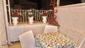 A small outdoor seating area with white chairs and a table covered with a cactus-patterned tablecloth. There are potted plants with red flowers on the windowsill, and a white railing with string lights.