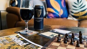 Et skakbræt, et glas mørk øl og en Guinness-dåse på et bord i et rum med blå vægge og en sofa.