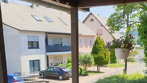 Een overdekt terras met een glazen tafel en stoelen, een houten schutting, en uitzicht op een straat met gebouwen en geparkeerde auto's.