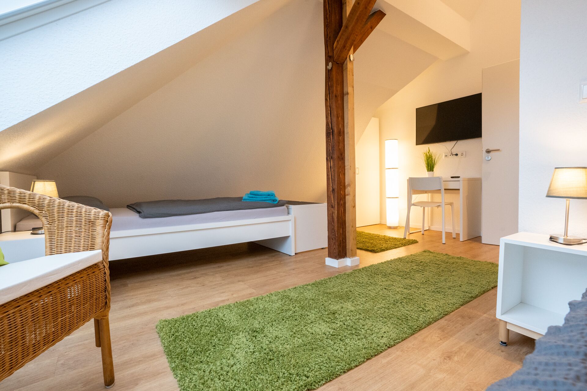 A loft bedroom with a sloped ceiling, a bed, a desk, a chair, and a TV mounted on the wall.
