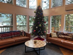 A cozy living room with a Christmas tree, leather sofas, and large windows overlooking a forest. - Lakefront Chalet w/ Hot Tub, EV Charger, King bed, Dock, Dogs OK! (Leavenworth)