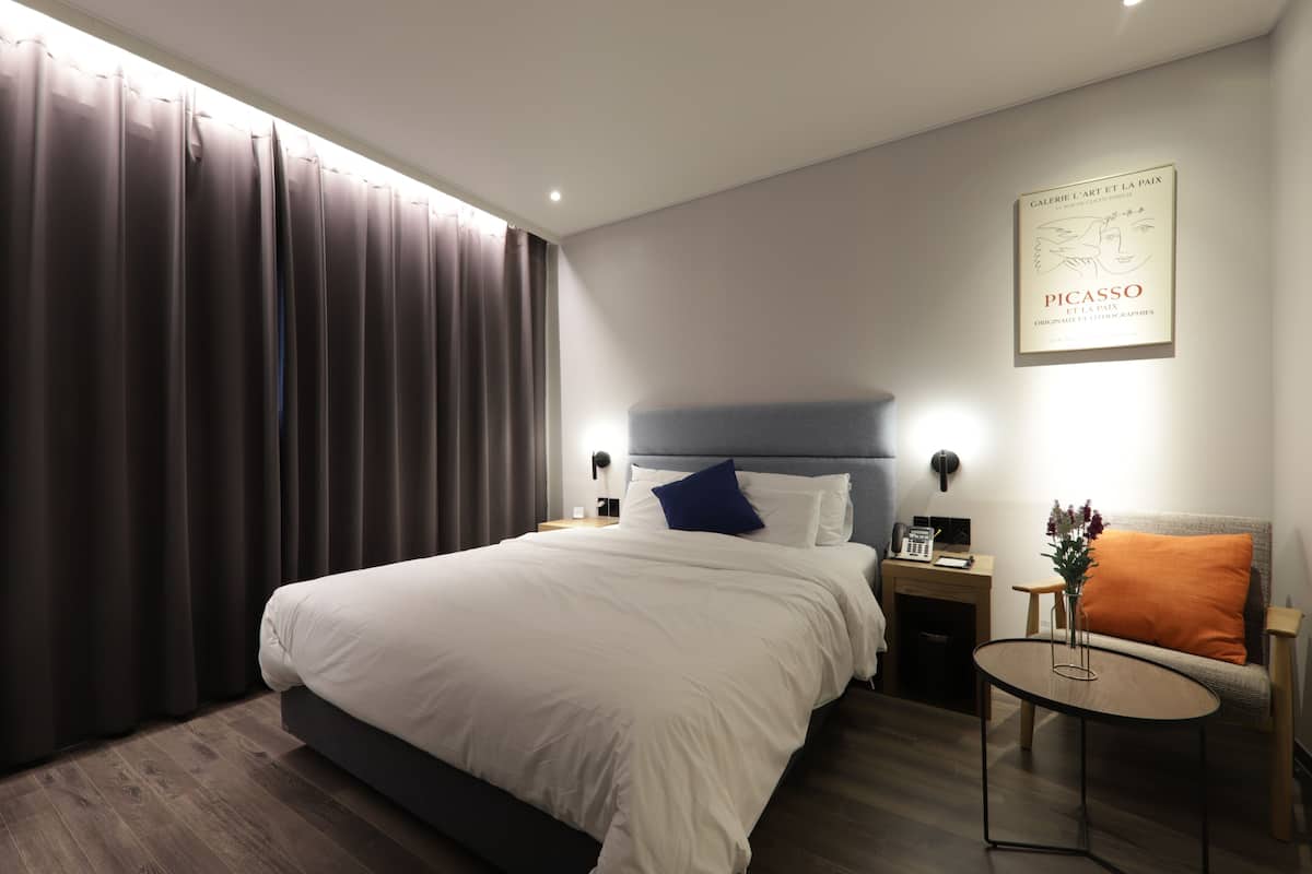 A modern hotel room with a large bed, a bedside table, a small round table, and a sofa.