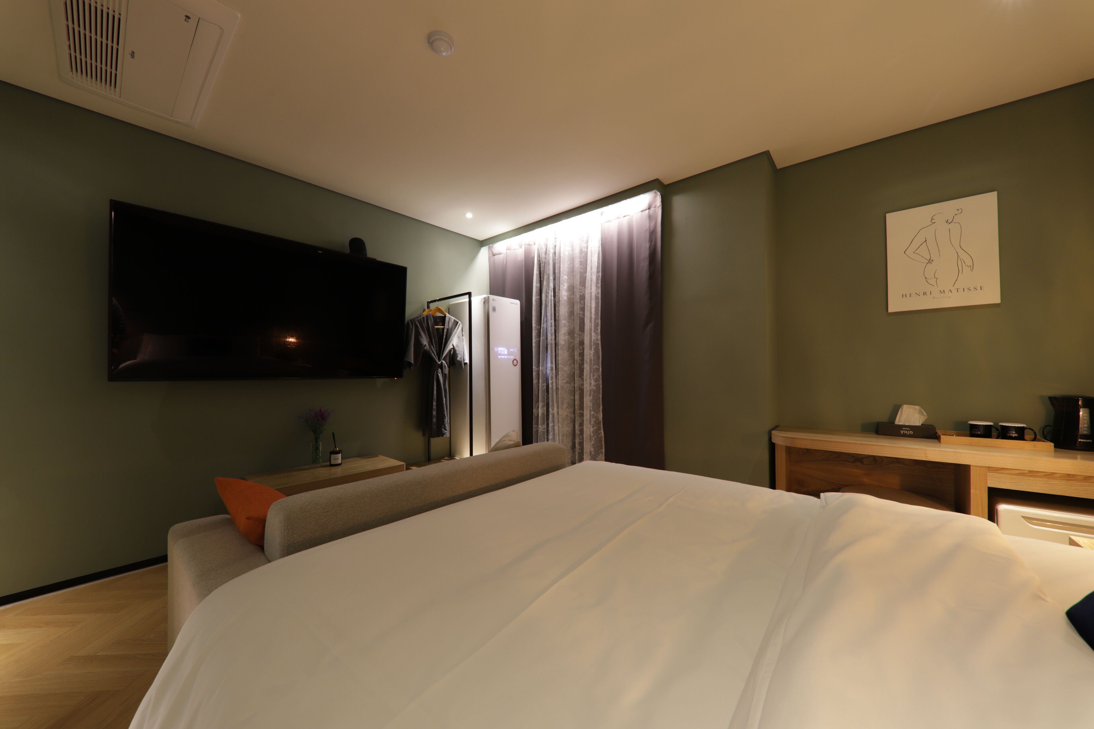 A modern hotel room with a large bed, a flat-screen TV mounted on the wall, a desk with a coffee maker, and a painting on the wall.