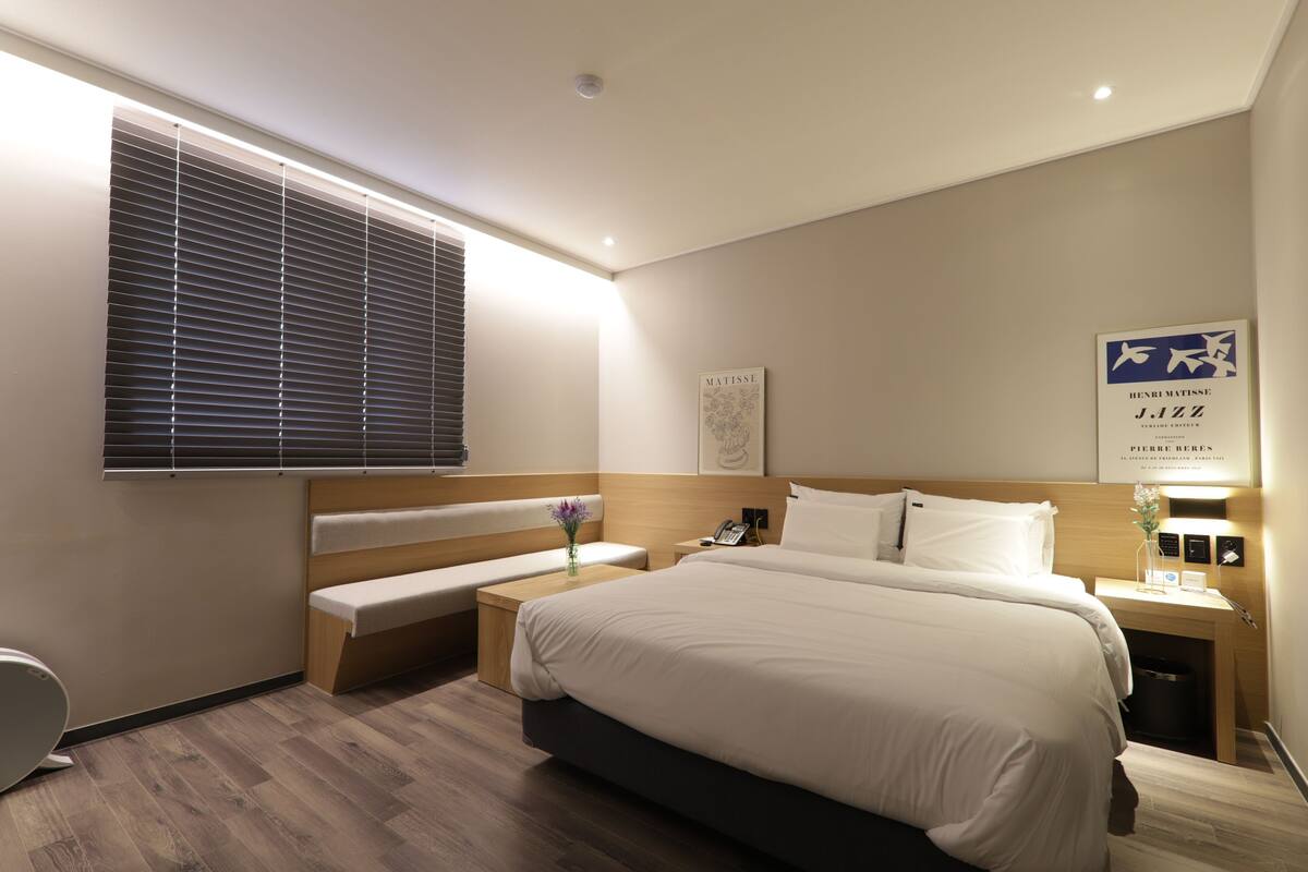 A modern hotel room with a large bed, bedside tables, a bench, a window with blinds, and artwork on the wall.