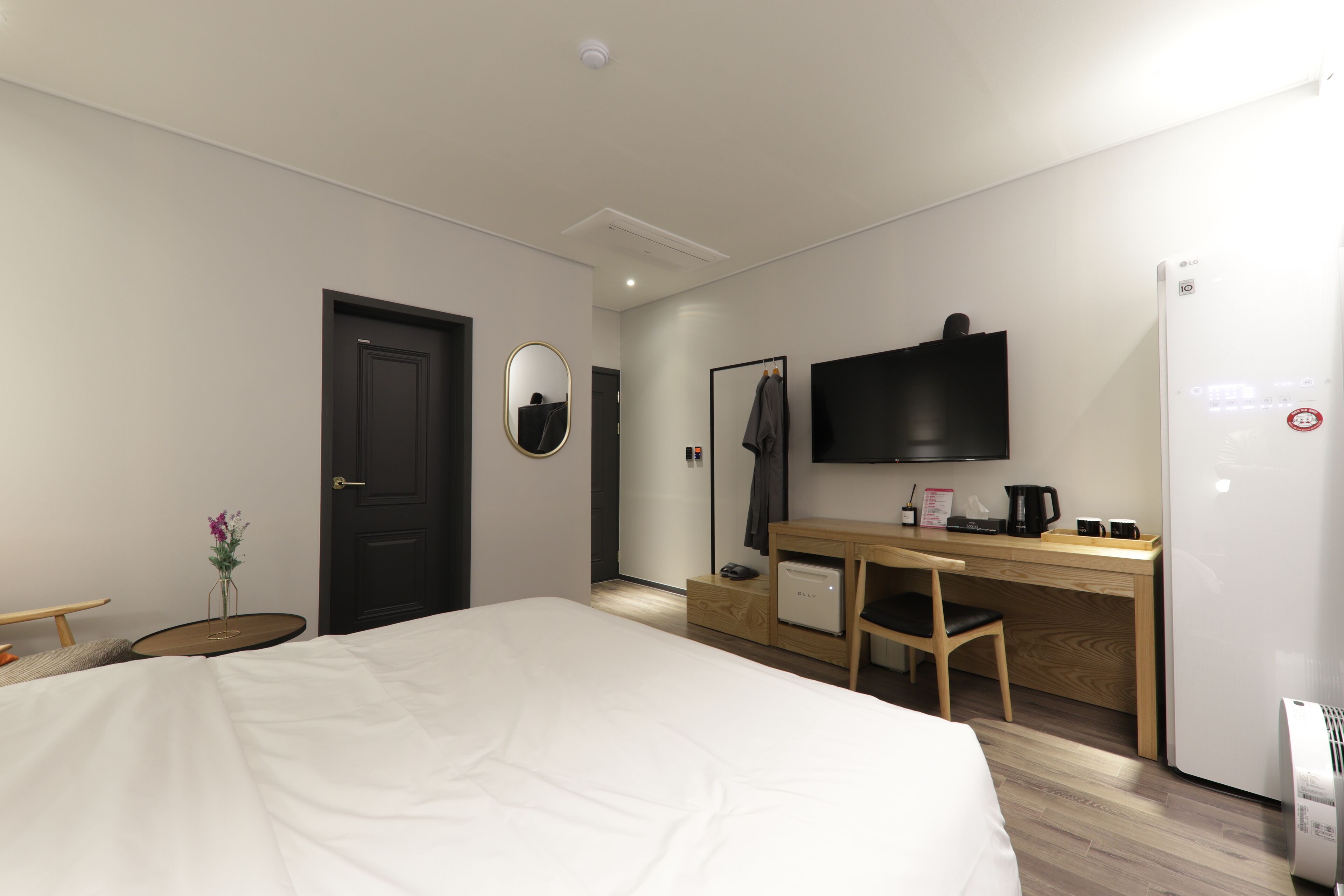 A modern hotel room with a large bed, a desk with a chair, a flat-screen TV mounted on the wall, a refrigerator, and a small table with a vase of flowers.