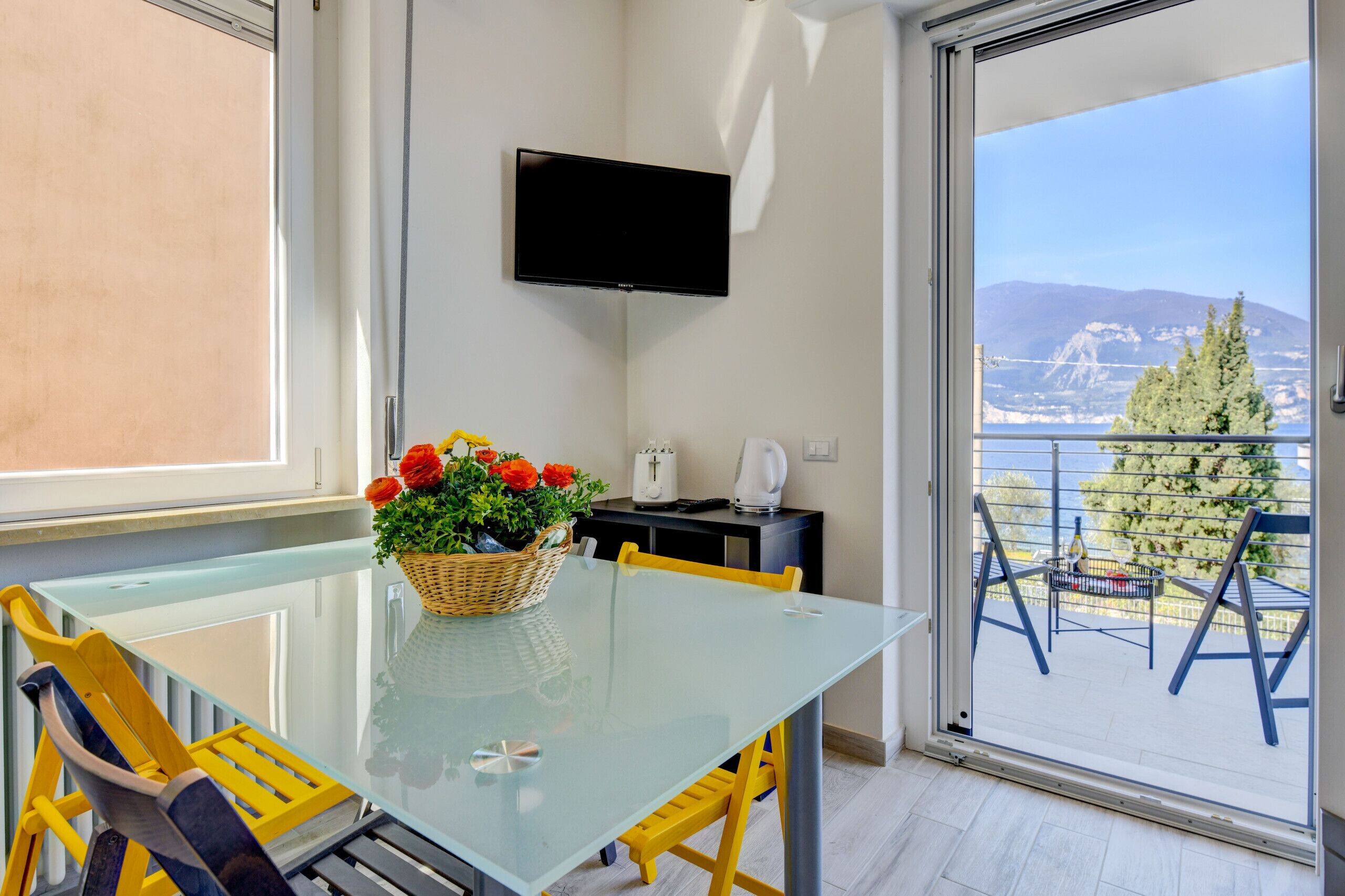A modern room with a glass table, yellow chairs, a flat-screen TV mounted on the wall, and a balcony with a view of the sea and mountains.