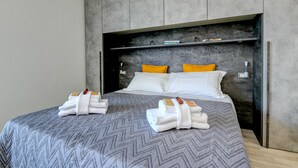 Une chambre d’hôtel moderne avec un grand lit, une couvre-lit grise à motifs, deux oreillers blancs et deux coussins décoratifs jaunes. On trouve deux jeux de serviettes blanches sur le lit.