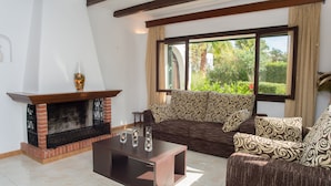 A living room with a fireplace, patterned sofa, and a large window with a view of greenery. - Spacious holiday home Dos Pinos with pool, A/C 24h, Wi-Fi, near Cala Esmeralda (Santanyí)
