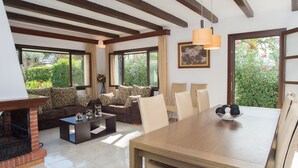 A living room with a fireplace, a sofa set, a dining table, and large windows with a view of greenery. - Spacious holiday home Dos Pinos with pool, A/C 24h, Wi-Fi, near Cala Esmeralda (Santanyí)
