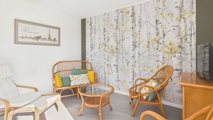 A living room with wicker furniture, a glass coffee table, a framed cityscape picture, and a wall with a nature-themed wallpaper. - Holiday home for 6 in Saint Hilaire de Riez (Saint-Hilaire-de-Riez)