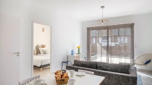 Ein modernes, gut beleuchtetes Zimmer mit einem Bett, einem Sofa, einem Esstisch mit Frühstücksgebäck und einem großen Fenster mit Blick auf Gebäude.