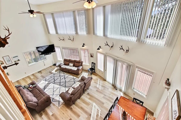 A spacious living area with a high ceiling, large windows, a ceiling fan, and a flat-screen TV.