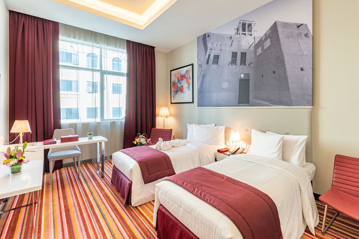 A hotel room with two beds, a desk, a chair, a lamp, and a large window with curtains.
