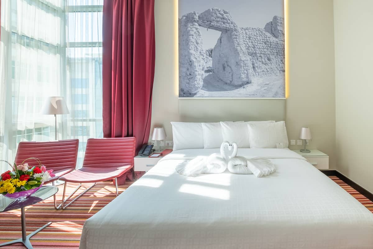 A hotel room with a large bed, a desk with a lamp, a chair, and a window with curtains.