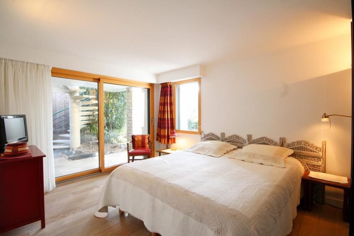 Une chambre avec un grand lit, un téléviseur posé sur un meuble, une chaise, une lampe et une vue sur un escalier et la verdure à l’extérieur.