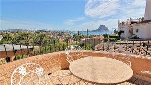 Terraço com mesa redonda e cadeiras de metal branco, com vista para o horizonte costeiro, incluindo uma montanha imponente e uma extensão de água.