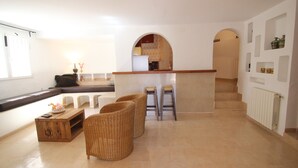Uma sala de estar espaçosa com cozinha ao fundo, apresentando uma mesa de centro de madeira, cadeiras de vime e uma geladeira branca.