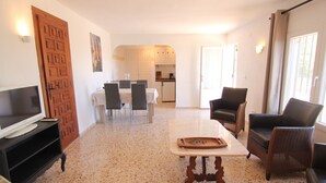 Uma sala de estar com TV, área de refeições com cadeiras, cozinha ao fundo e uma janela com cortinas.
