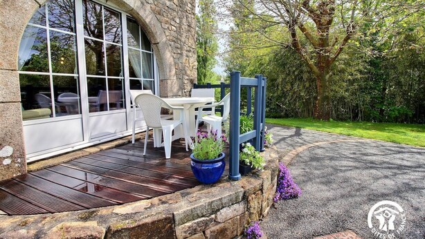 A stone building with a large window, a patio with a table and chairs, and a garden with trees and plants. - Pont Bihan - CARNAC - Gîte (CARNAC)