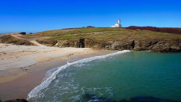Eine Küstenlandschaft mit einem Leuchtturm auf einem grasbewachsen Hügel, einem Sandstrand und ruhigem Meer.