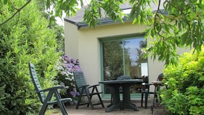 Un patio al aire libre con una mesa y sillas, rodeado de abundante vegetación y un edificio con un ventanal grande.